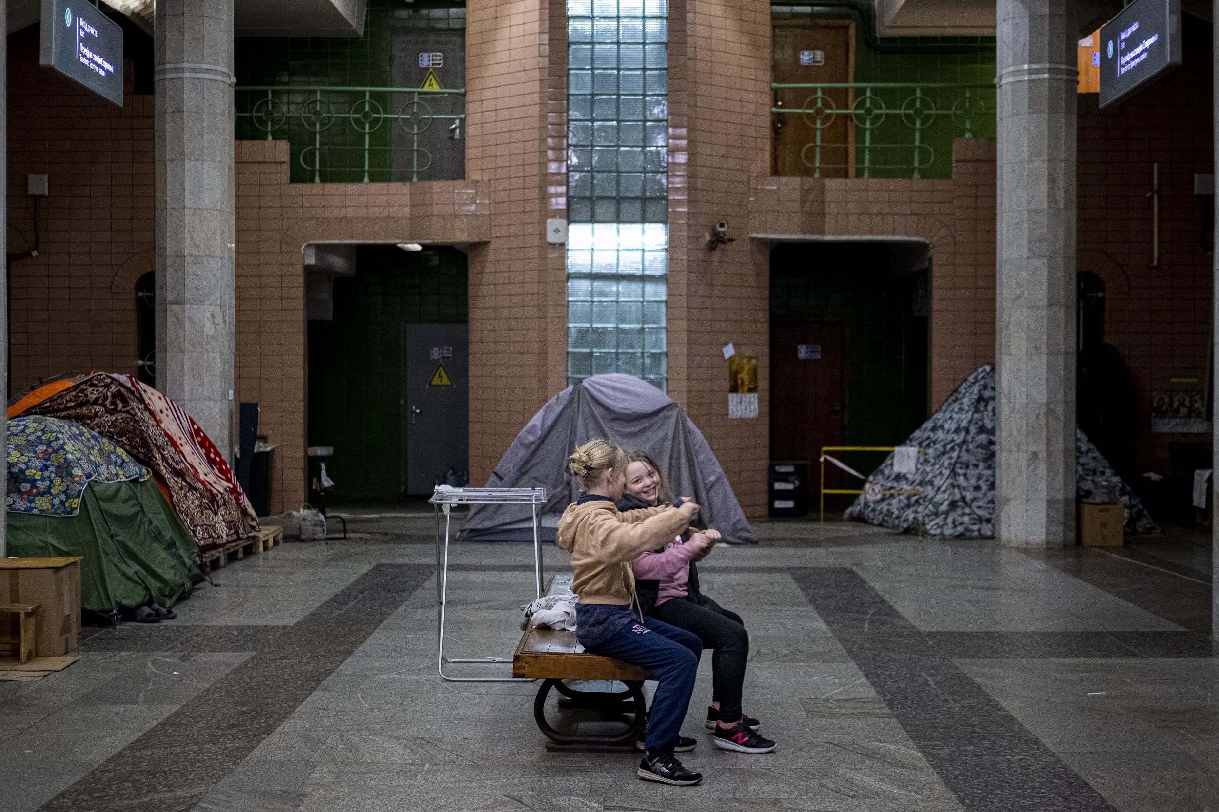 Two girls smile as they sit on a bench in a subway station.